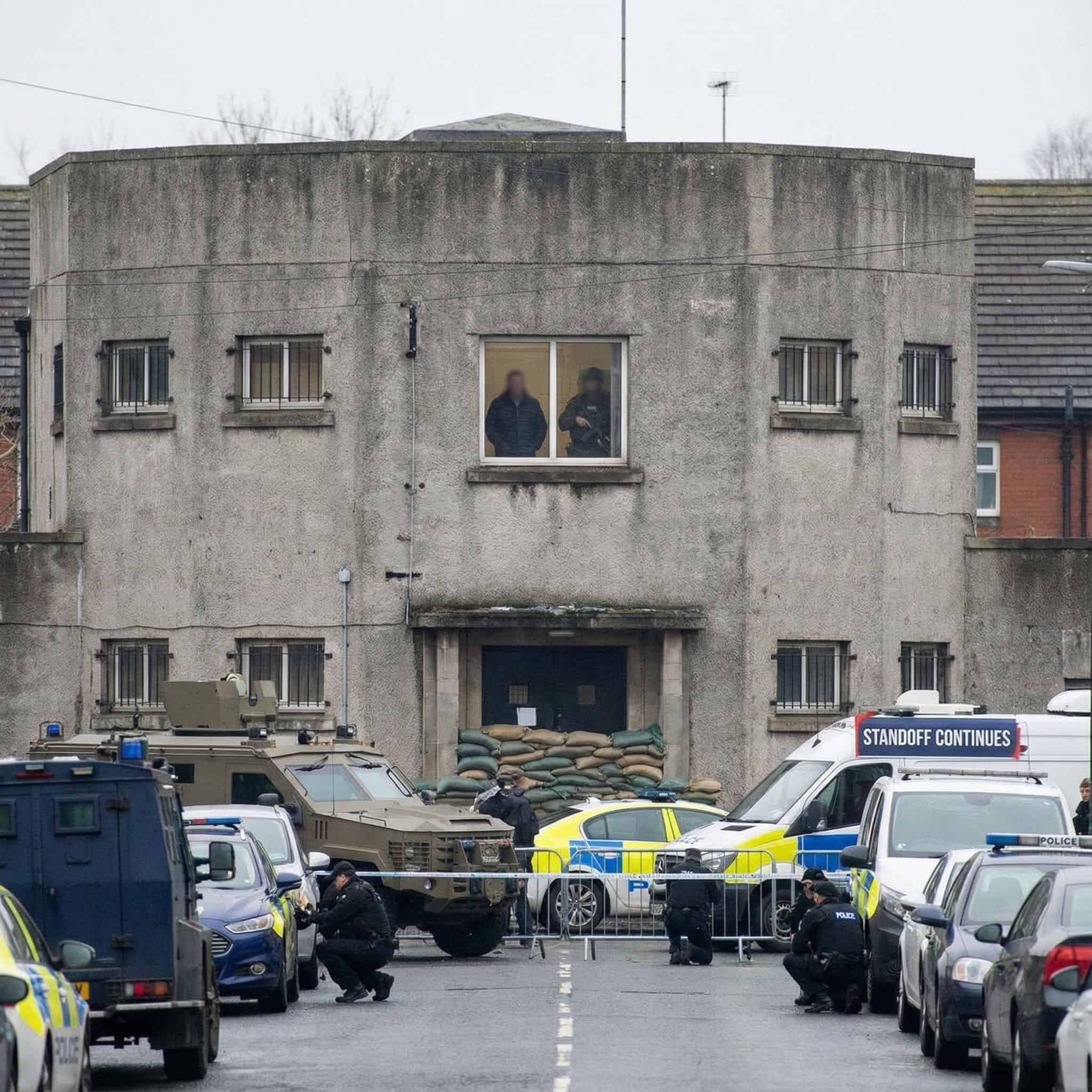 인질이 있는 요새화된 건물 안쪽으로, 창문과 문을 막고 방어 태세를 취한 무장 테러리스트들의 긴장된 대치 상황을 보여준다.